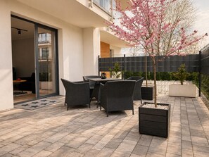 Une terrasse avec des meubles d’extérieur, un petit arbre aux fleurs roses et une clôture noire.