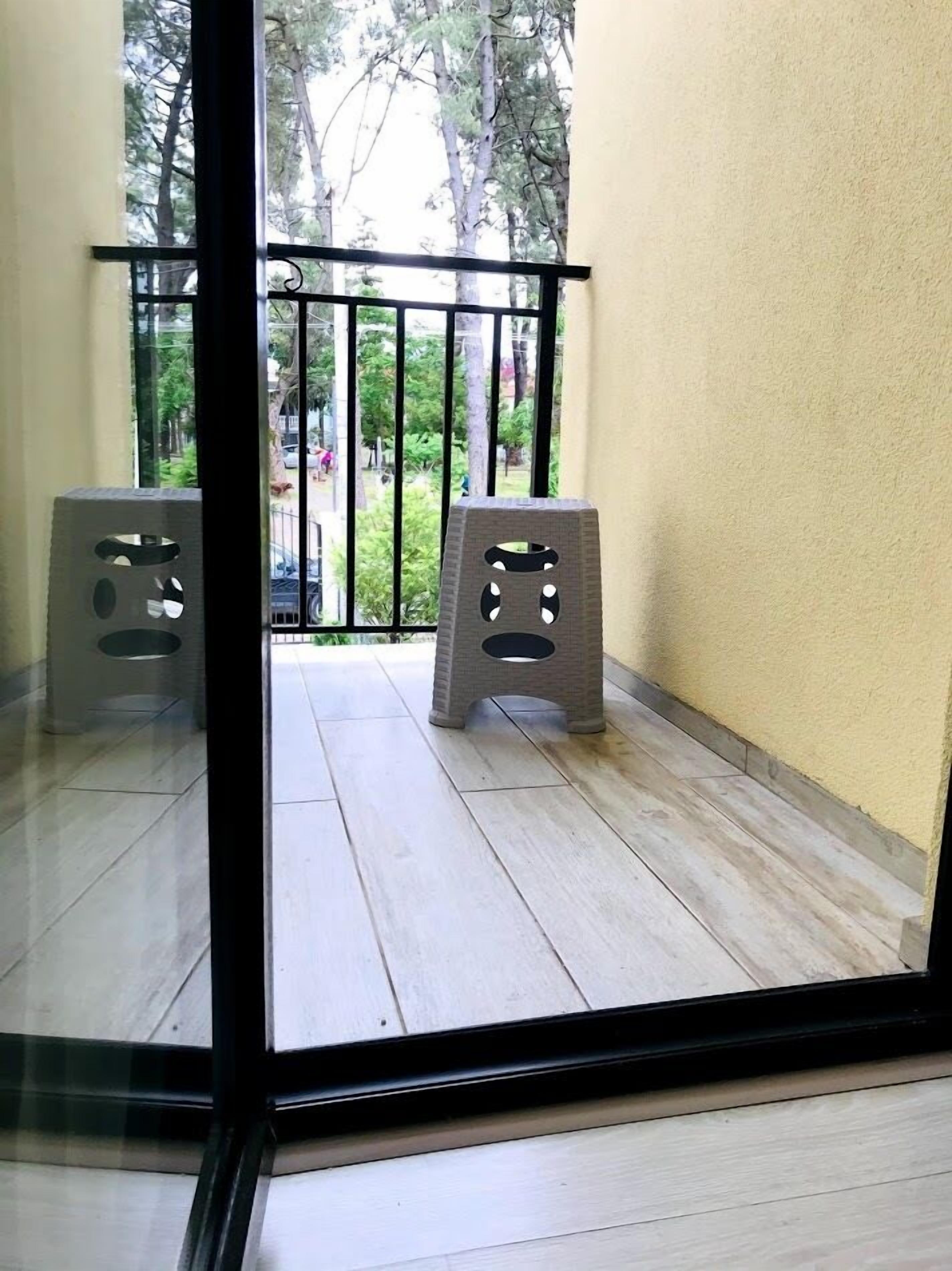 Un balcón con puerta de cristal, dos taburetes y vista a la calle y árboles.
