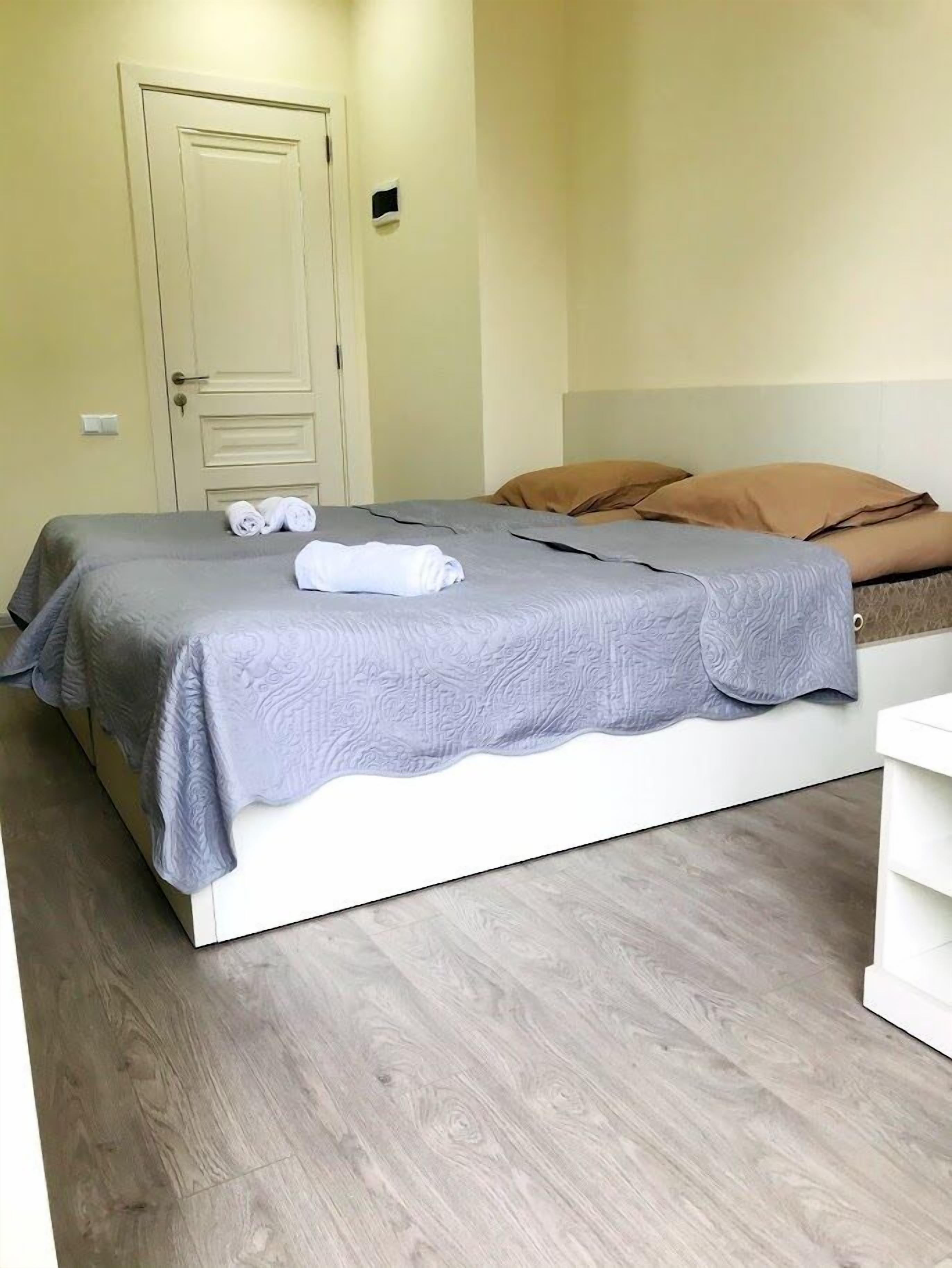 A neatly made bed with a grey bedspread and rolled towels, a white bedside table, and a wooden floor.