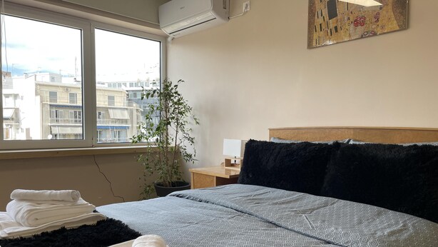 A hotel room with a bed, a potted plant, a window with a view of buildings, and a painting on the wall. - Cozy Apartment in Heart Athens Near Metro Fix 100m (Athens)