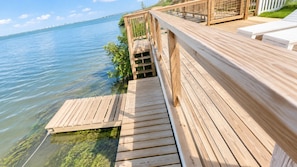 A wooden deck extending over water with a railing, a clear view of the water, and a distant bridge.
