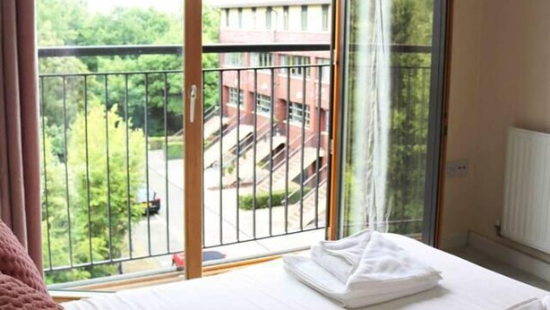 Ein Hotelzimmer mit einem Bett, einer rosa Decke und Blick auf ein Gebäude und Grün durch einen Balkon mit Metallgeländer.