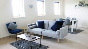 Ein Wohnzimmer mit grauem Sofa, blauem Sessel und einem Couchtisch. Im Hintergrund steht ein Esstisch mit Stühlen.