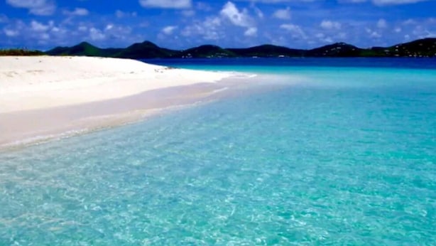 Una playa con agua turquesa cristalina y arena blanca, rodeada de montañas.