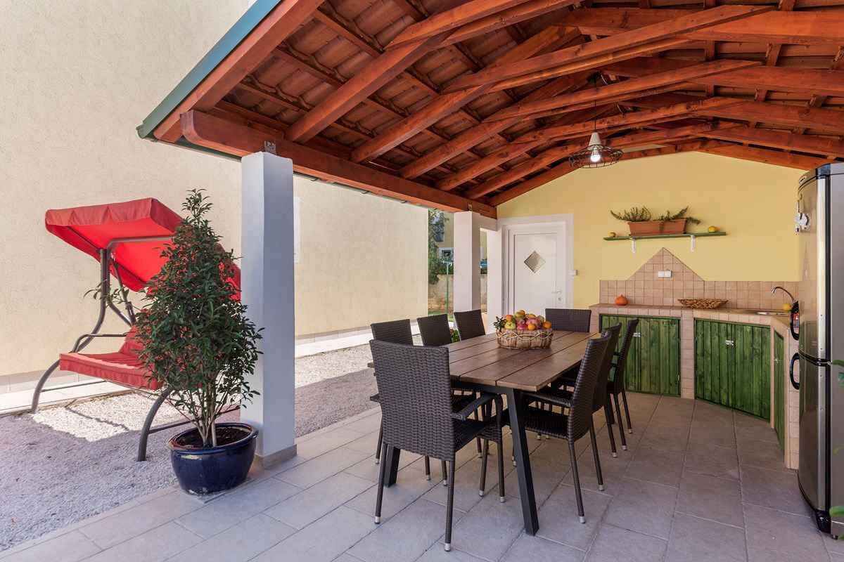 Eine Terrasse im Freien mit Holzboden, einem Esstisch mit Stühlen, einem Blumentopf und einem roten Sonnenschirm.