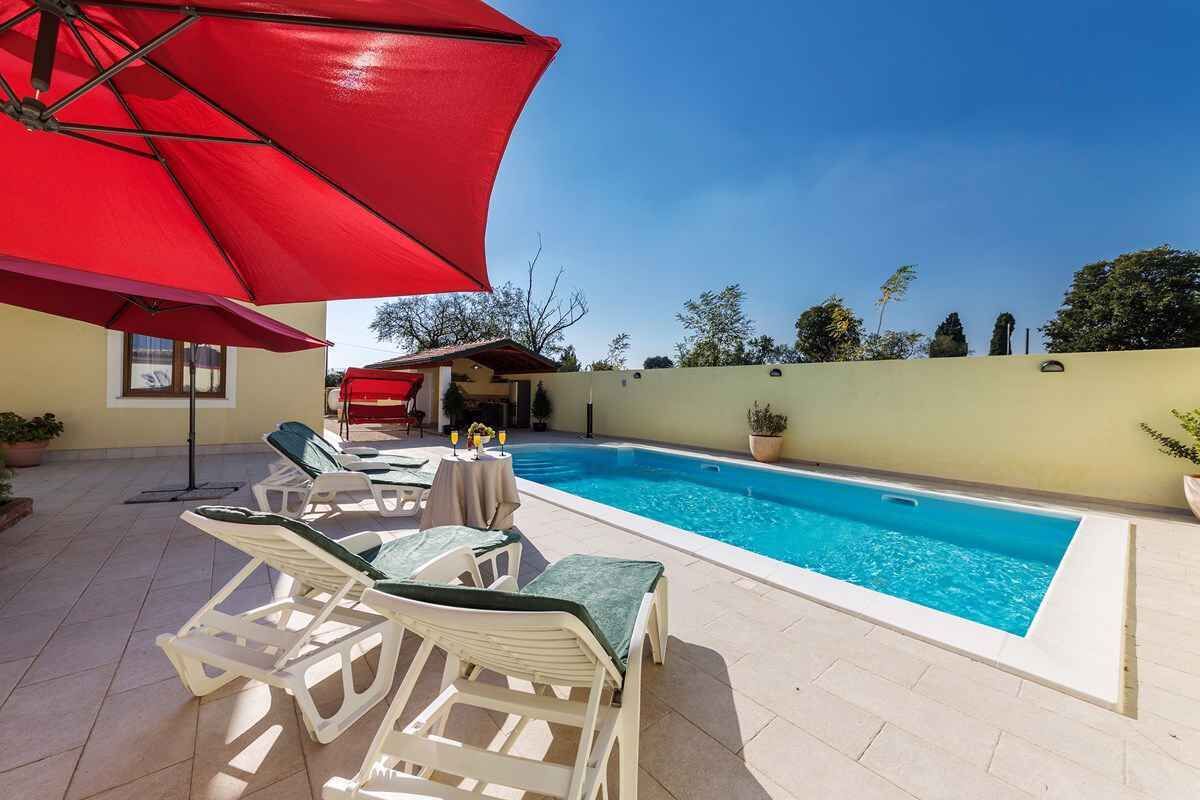 Une piscine entourée d’une terrasse carrelée, avec des chaises longues et des parasols rouges.