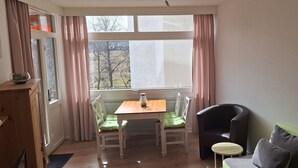 Um quarto com uma mesa de madeira, duas cadeiras, uma mesinha redonda, um sofá verde e uma janela com cortinas.