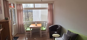 Une pièce comprenant une table en bois, deux chaises, une petite table ronde, un canapé vert et une fenêtre avec des rideaux.