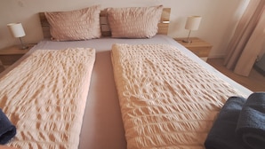 Cama de casal com duas almofadas, duas mesas de cabeceira com abajures e uma janela com cortinas.