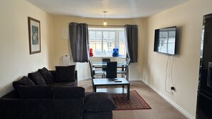 Uma sala de estar com um sofá de cor escura, uma mesa de jantar com cadeiras, uma TV fixada na parede e uma janela com cortinas.
