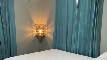 Et soveværelse med en seng, lyseblå gardiner, en vægmonteret lampe og en aircondition.