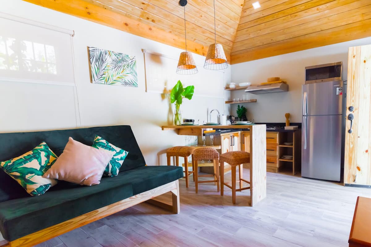 Un salon chaleureux avec un canapé vert, des chaises en bois et un coin cuisine équipé d’un réfrigérateur et de meubles de rangement en bois.