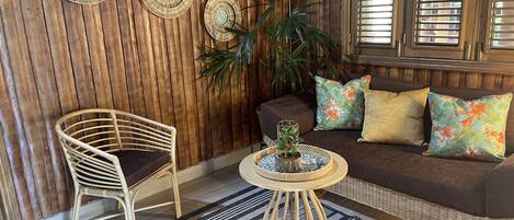 A room with a wooden wall, a wicker chair, a sofa with colorful cushions, a small table with a plant, and two wall decorations.