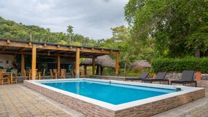 Una piscina rectangular con borde de ladrillo, rodeada de sillas de descanso y una pérgola de madera con techos de paja.