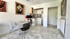 A modern kitchen with white cabinets, a microwave, and a refrigerator. There is a dining area with a table and chairs, and a painting on the wall. - Bright 31m2, flowered terrace, near Croisette (Cannes)