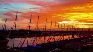 Un puerto deportivo con varios veleros amarrados, un cuerpo de agua tranquilo y un cielo al atardecer.