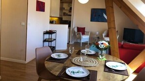 Comedor con mesa redonda dispuesta para cuatro, cocina al fondo y sala de estar con televisor y sofá rojo.