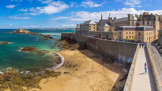 Un pueblo costero con un imponente edificio parecido a un castillo, una playa de arena y un cielo azul despejado.