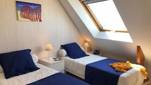 Una habitación pequeña y bien iluminada con dos camas, cada una con una colcha azul y almohadas blancas. Hay un cuadro en la pared, una ventana con vistas, y una mesita con una lámpara y un bolso.