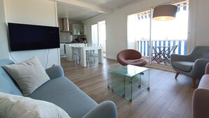 A modern living room with a sofa, a glass coffee table, and a TV mounted on the wall. There is a dining area with a table and chairs, and a balcony with a railing.