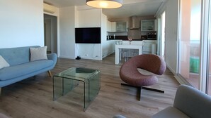A modern living room with a sofa, a glass coffee table, and a unique curved chair. A flat-screen TV is mounted on the wall, and a kitchen is visible in the background.