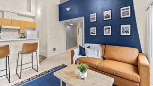 A modern living room with a blue accent wall, a brown leather sofa, two bar stools, a coffee table with a plant, and a kitchen area with wooden cabinets and a white refrigerator. - Emerald City Pentouse (Seattle)