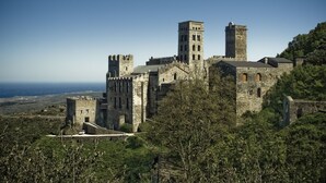 Un castillo histórico con múltiples torres y una muralla fortificada, rodeado de vegetación exuberante y con vistas a un paisaje costero.