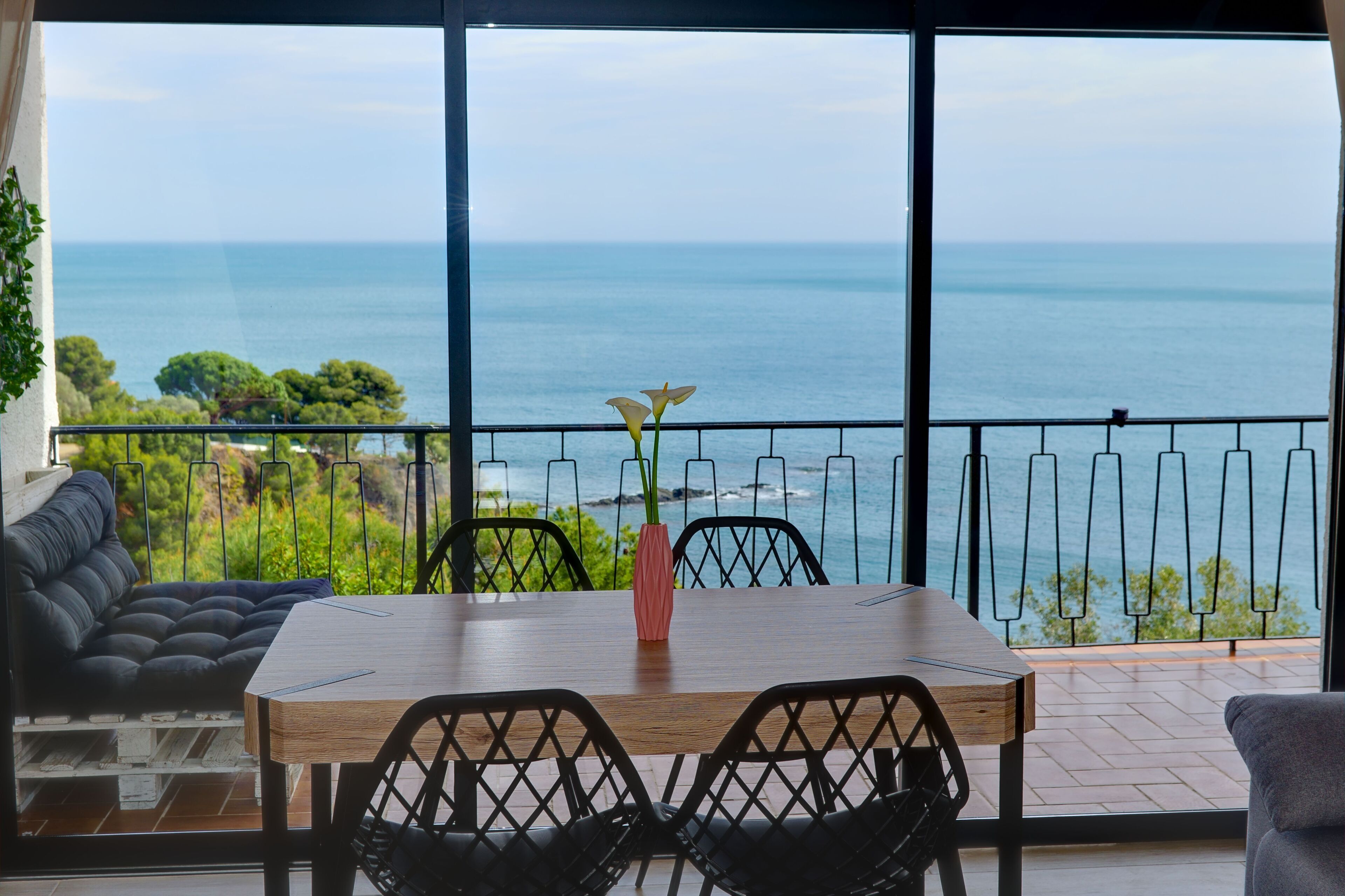 Uma sala de jantar com uma mesa e cadeiras de madeira, um vaso rosa e vista para o oceano.