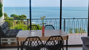 Un comedor con una mesa y sillas de madera, un florero rosa y vista al océano.