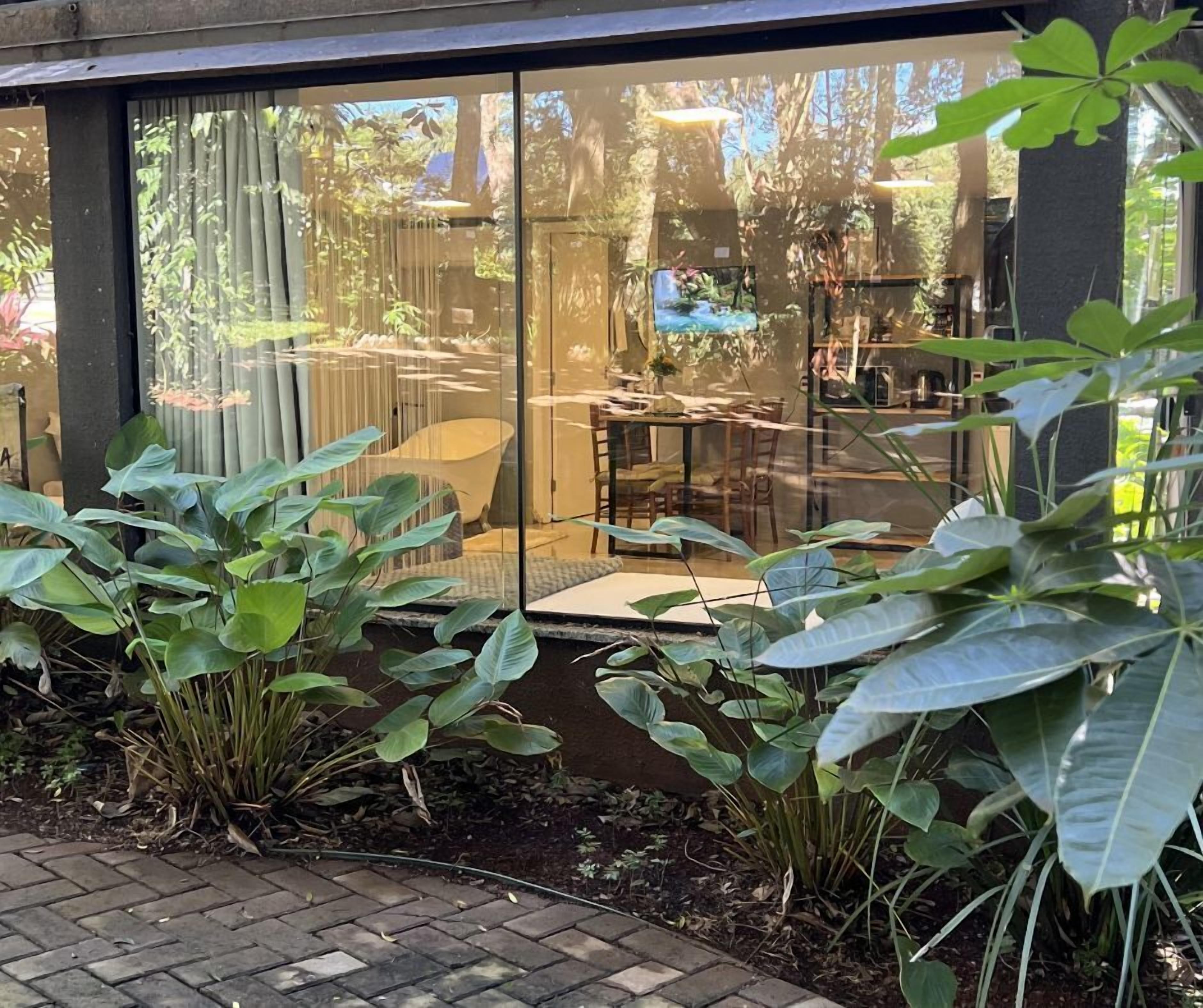 Un espacio moderno para comer al aire libre con cerramiento de vidrio, rodeado de abundante vegetación y un sendero de ladrillo.