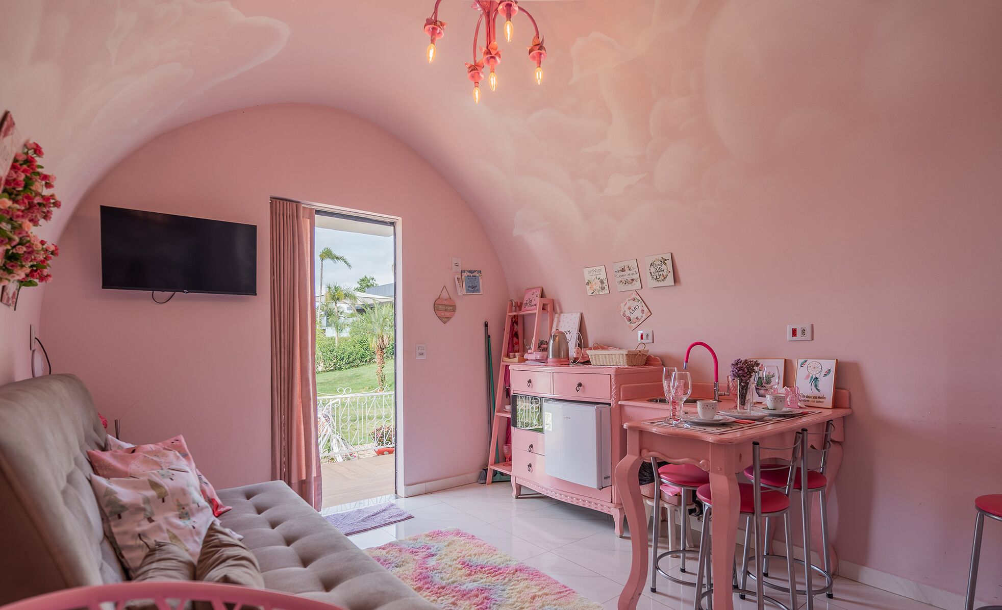 A pink-themed room with a sofa, a desk with a chair, and a TV mounted on the wall.