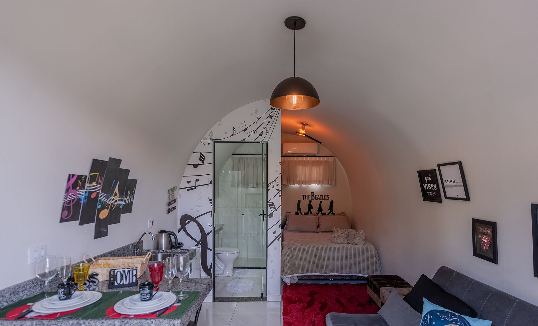A compact living space with a kitchenette, a dining area, a bedroom with a red rug, and a wall with musical notes and a Beatles poster.