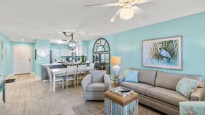 Un salon comprenant un canapé, un fauteuil et une table basse, un coin repas avec une table et des chaises, et une cuisine équipée de placards et d’appareils électroménagers.