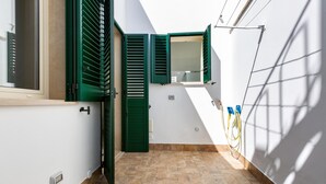 A narrow outdoor corridor with a tiled floor, green shutters, a small window, and a wall-mounted electrical outlet.