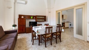 A dining area with a table set for four, wooden chairs, a sofa, and a kitchen in the background.