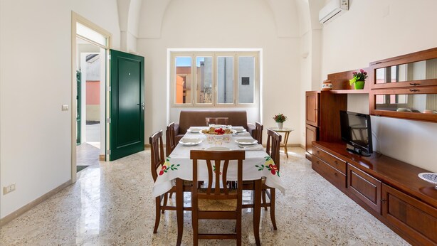 A dining area with a table set for four, flanked by wooden chairs, a television on a wooden cabinet, and a window with a view of buildings outside.