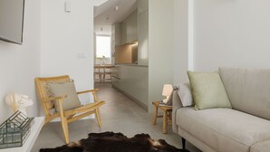 Un salon moderne avec un canapé, une chaise en bois et une petite table. On aperçoit une cuisine au fond.