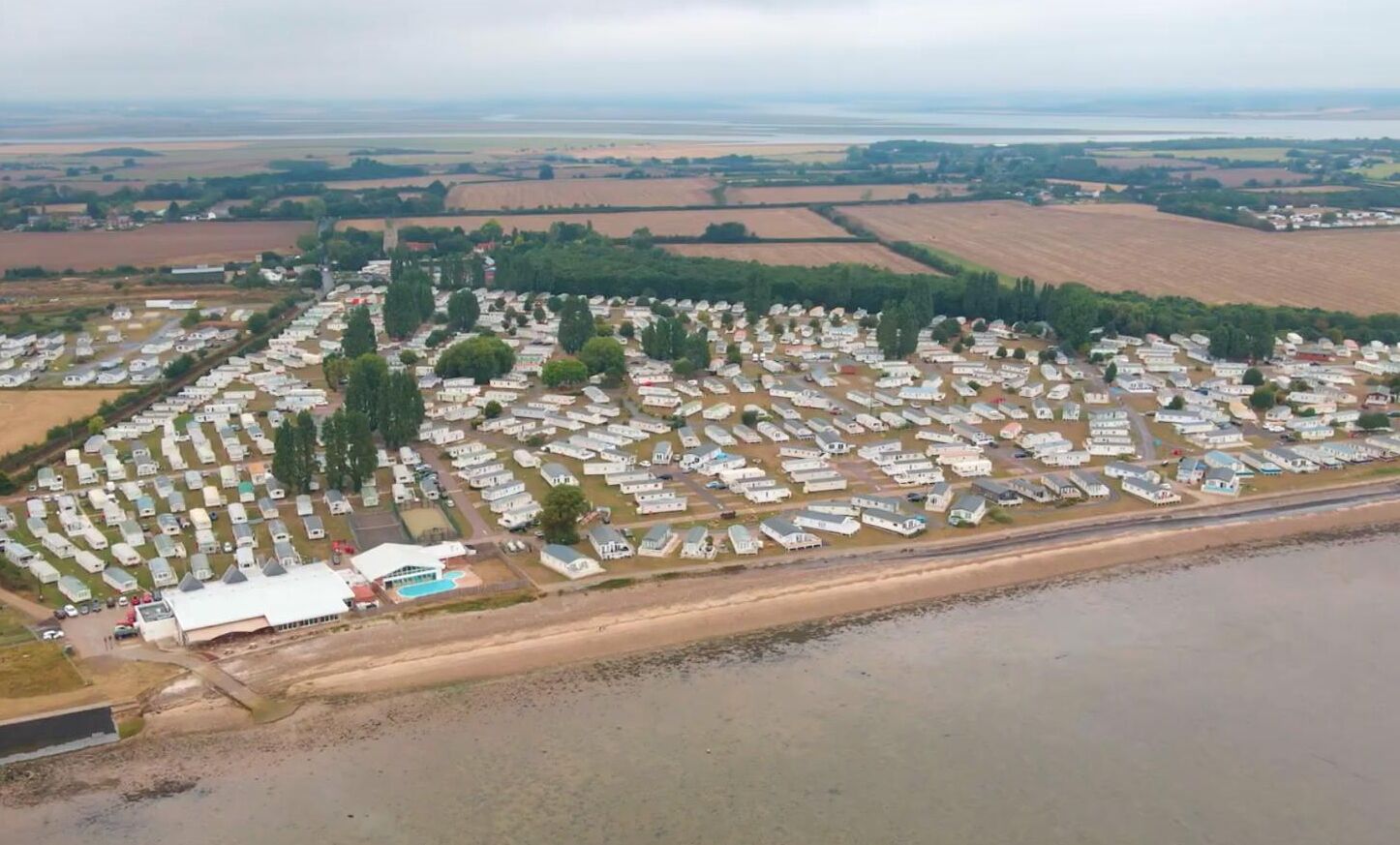 An aerial view of a campground with numerous RVs and trailers arranged in rows, a beach in the foreground, and fields in the background.