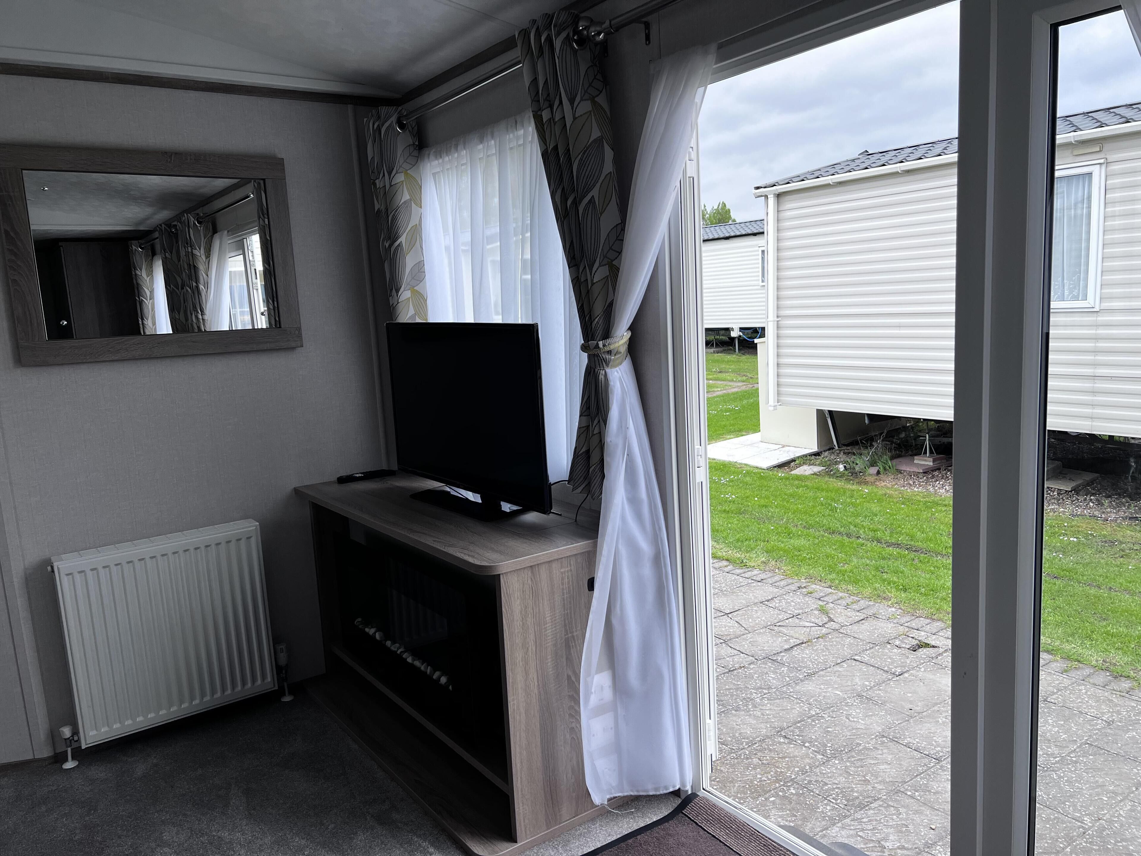 A room with a television on a wooden stand, a radiator, and a view of a grassy yard and a mobile home.