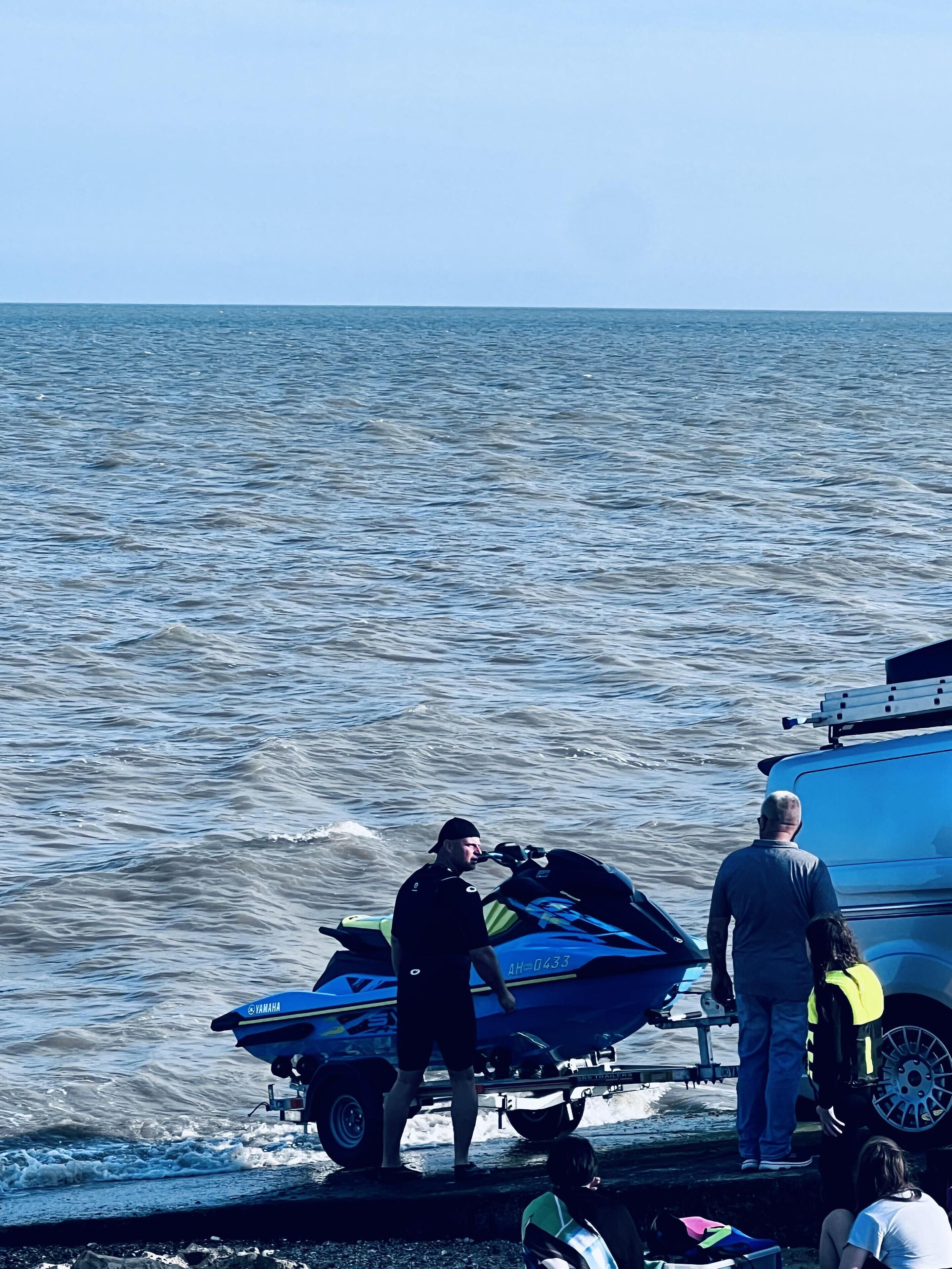 Ein Jetski auf einem Anhänger am Wasser, ein Boot im Hintergrund und Personen in der Nähe.