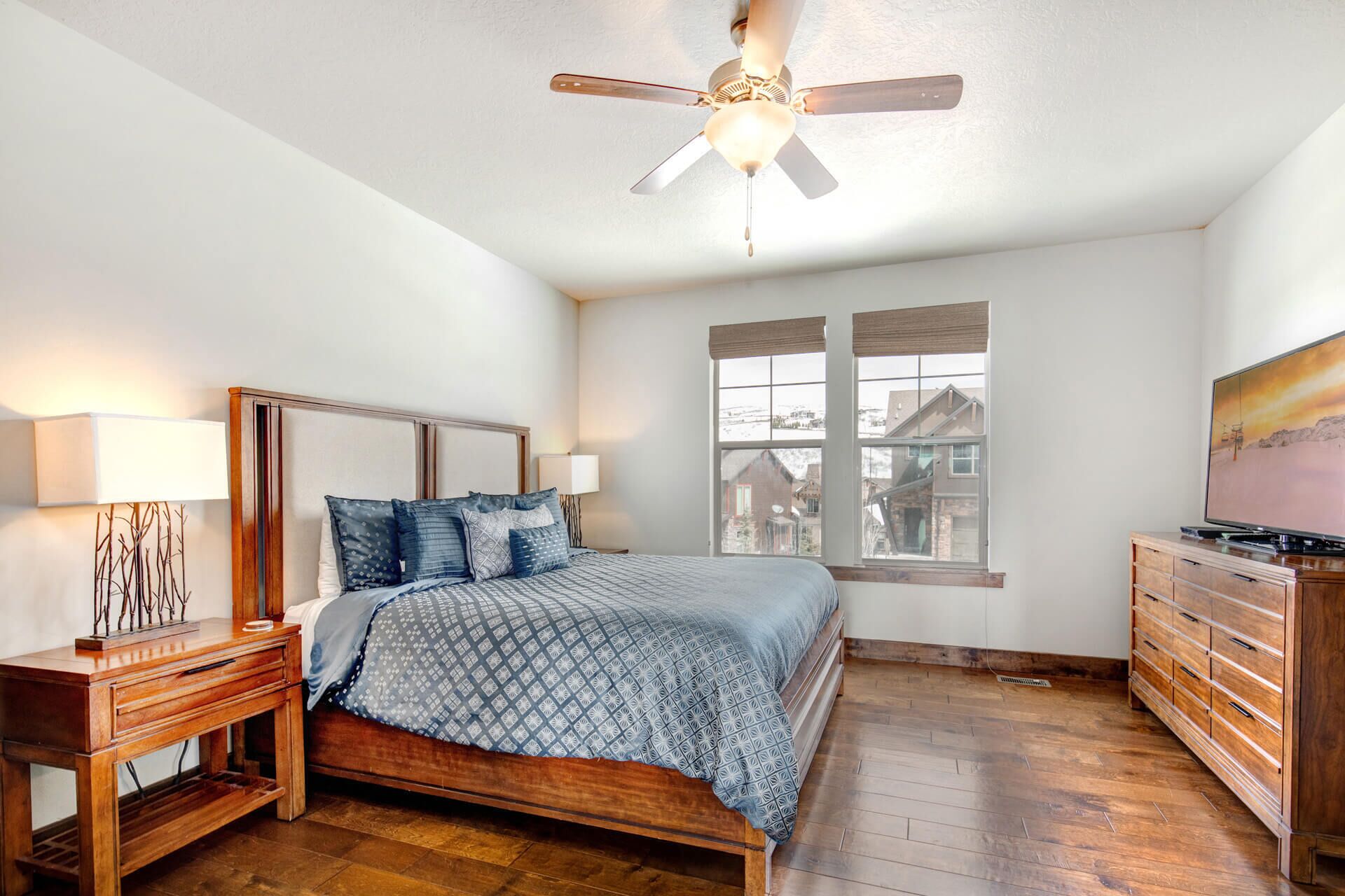 Une chambre à coucher avec un lit en bois, une table de chevet, une commode, un ventilateur de plafond et une fenêtre donnant sur des maisons.