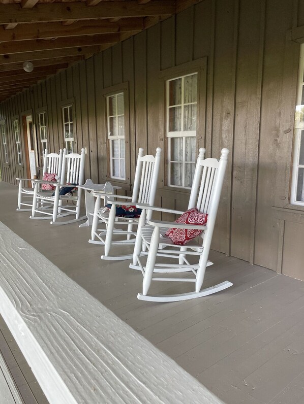A covered porch with white rocking chairs and a bench, wooden walls, and windows with curtains.