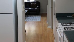 Kapea keittiö, jossa on jääkaappi, liesi ja kaappeja. Taustalla näkyy ruokailutila, jossa on sohva ja matto.
