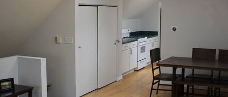 Uma área de refeições com mesa e cadeiras de madeira, ventilador de teto e cozinha ao fundo.