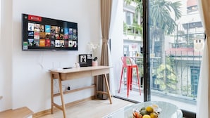 A modern living room with a large TV displaying a Netflix interface, a wooden desk, a glass coffee table with fruit, and a view of a balcony with plants and buildings. - Margaery Boutique Apartment (Hanoi)