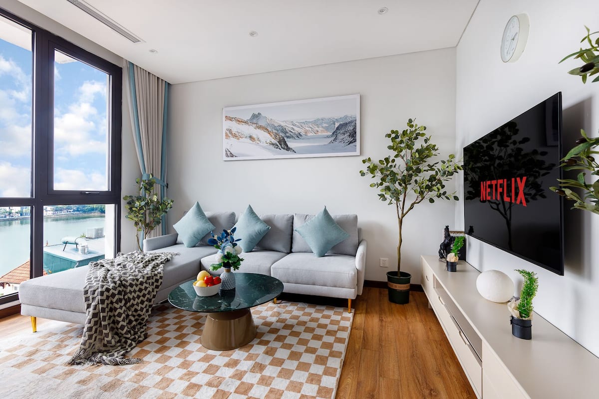 Een moderne woonkamer met een bank, salontafel en planten, een groot raam met uitzicht en een flatscreen televisie aan de muur.