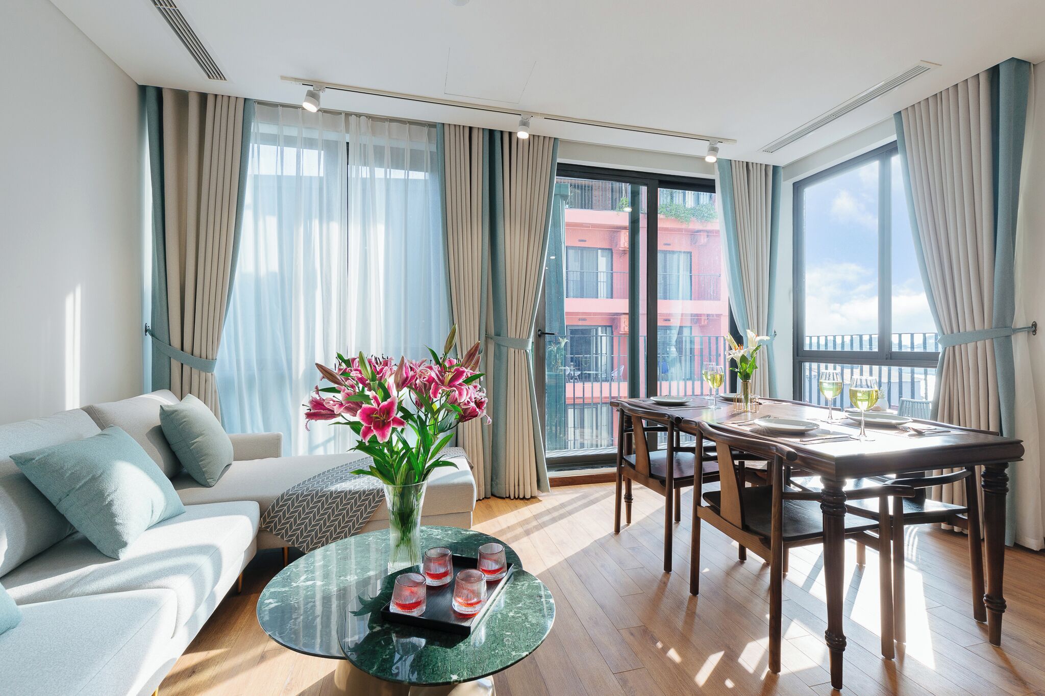 A modern living room with a sofa, a glass coffee table with decorative items, a dining table set for two, and large windows with curtains.