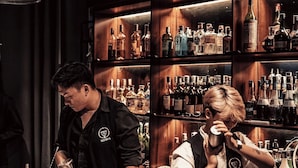 A bar with shelves stocked with various bottles, a bartender preparing drinks, and a customer in the background. - Margaery Boutique Apartment (Hanoi)