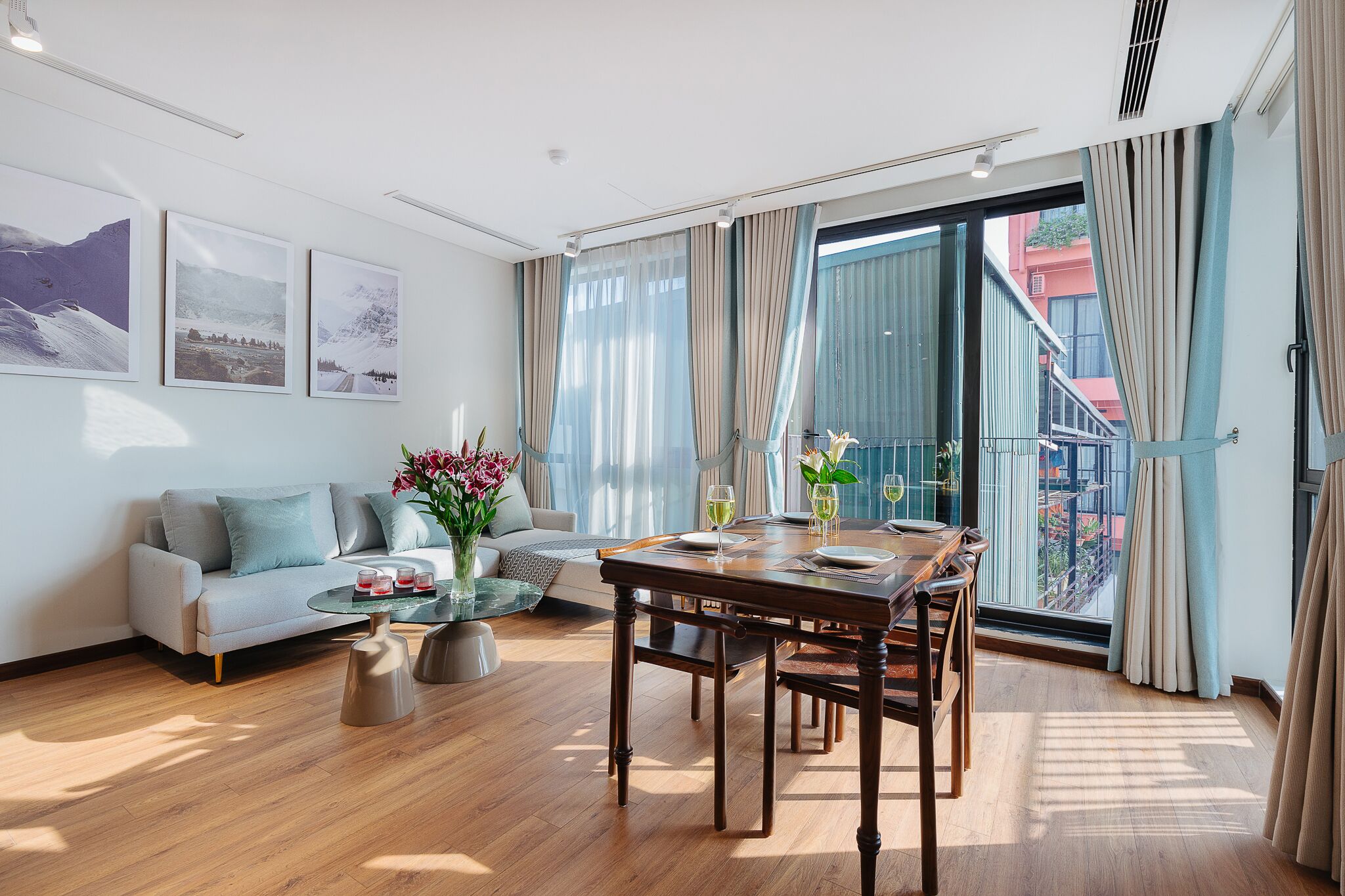 A modern living room with a sofa, coffee table, dining table, and chairs. There are large windows with curtains, a balcony, and artwork on the wall.