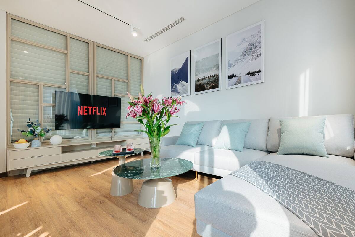 Een moderne woonkamer met een flatscreentelevisie, een glazen salontafel, een witte bank en een decoratieve vaas met bloemen.